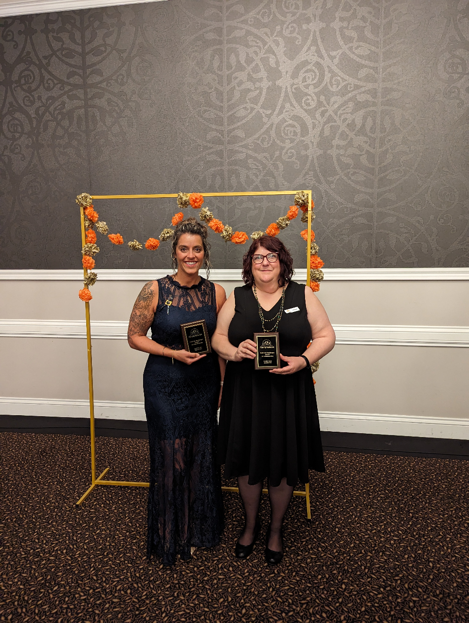 This is a photo of Jodi and Leslie, two esteemed individuals. Jodi, on the left, is dressed in a dignified dark grey lace gown, while Leslie, standing next to her, wears a black short-sleeved dress. They are positioned in front of a majestic gold arch adorned with vibrant orange and gold paper flowers. Each holds a plaque, The Matthew MacGregor Award, symbolizing their outstanding achievement.
